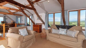 Un vaste salon doté de hauts plafonds et de poutres apparentes, deux canapés beiges ornés de coussins à motifs, une commode en bois et de grandes fenêtres offrant une vue sur la campagne.