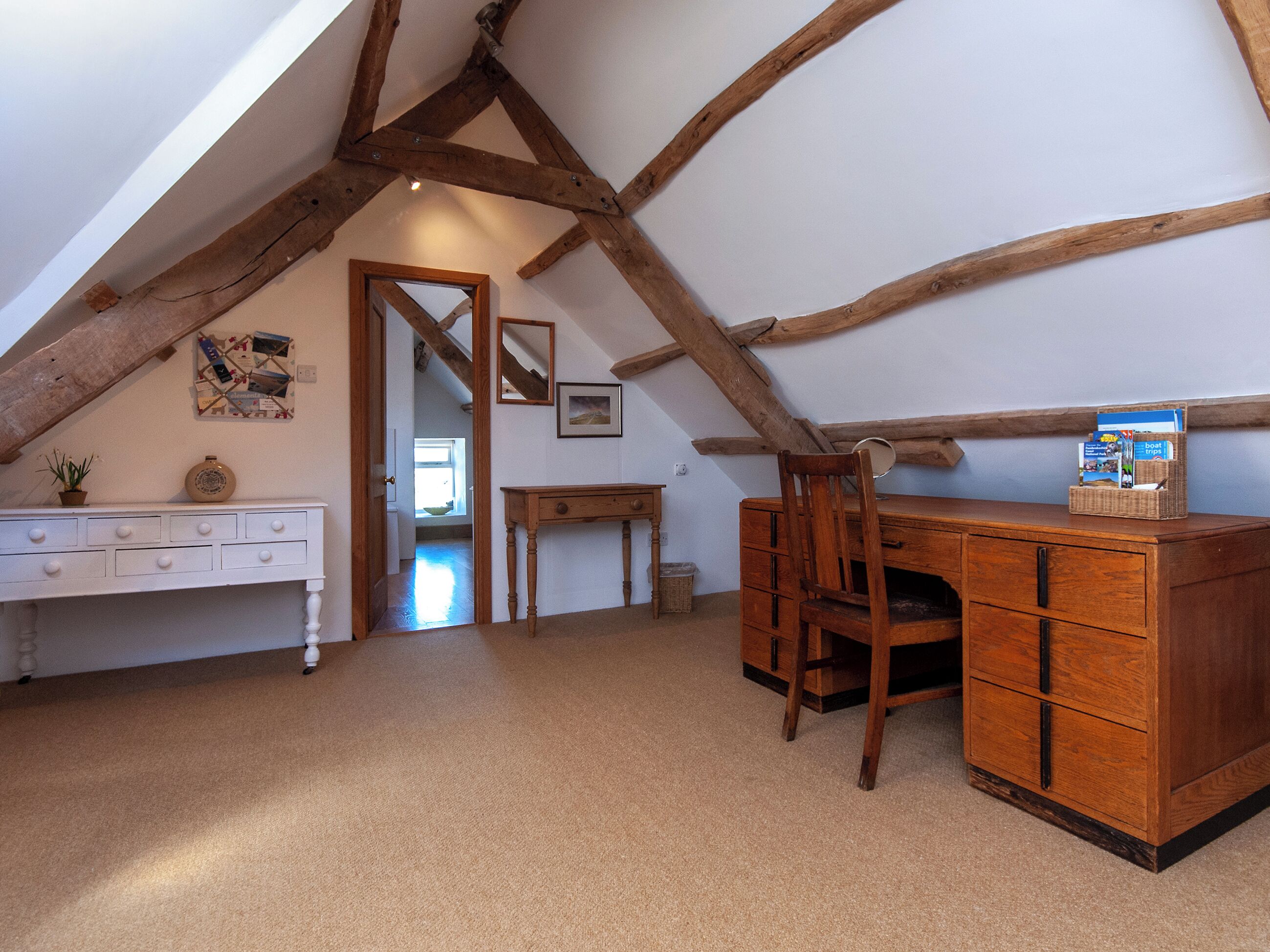 A room with a sloped ceiling, wooden beams, a desk, a chair, and a dresser. There is a window and a door leading to another room.