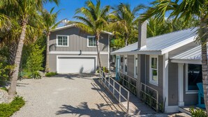 A two-story house with a garage, surrounded by palm trees and a gravel driveway. - Beach Don't Kill My Vibe by Beach Boutique Rentals (Anna Maria)