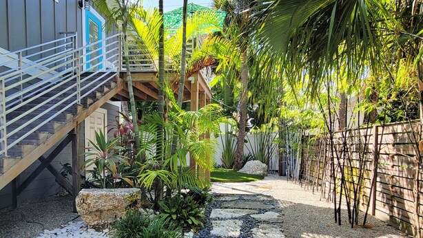 A tropical garden with a stone pathway, bamboo fence, and a staircase leading to a wooden deck. - Beach Don't Kill My Vibe by Beach Boutique Rentals (Anna Maria)