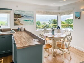 Une cuisine dotée d’un plan de travail en bois, d’une table à manger avec des chaises, et de grandes fenêtres offrant une vue sur la verdure et les montagnes.