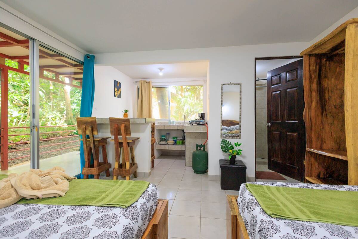 A room with two beds, a dining table, a kitchen area, and a sliding glass door to the outside.