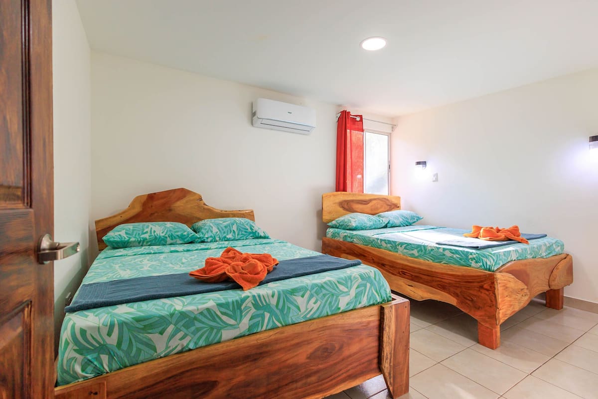 Two wooden beds with leaf-patterned bedding, a red curtain, and an air conditioning unit on the wall.