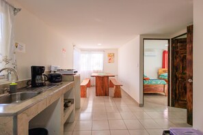 A compact kitchen with a sink, coffee maker, and a wooden table with benches. A bedroom with a bed and a red curtain is visible through an open door.