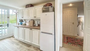 Una cocina con armarios blancos, un refrigerador, un microondas y una tabla de cortar de madera sobre la encimera.