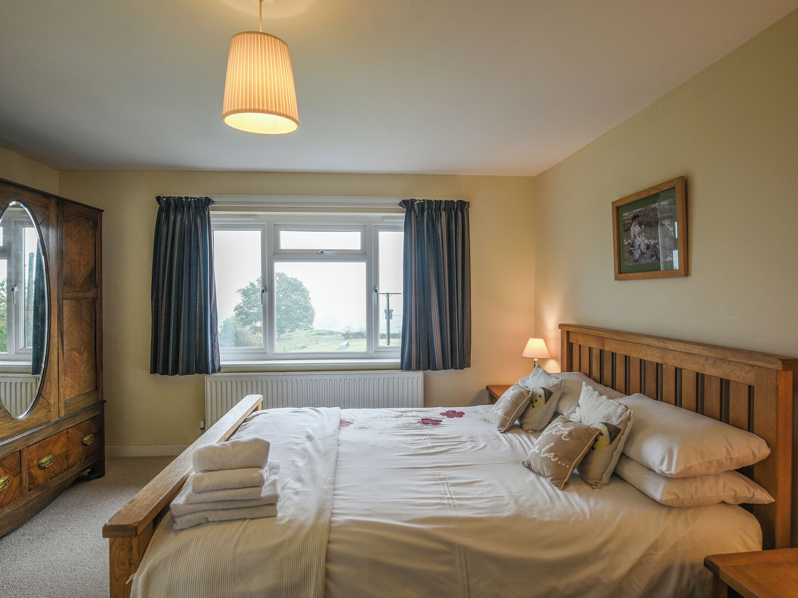 Une chambre à coucher avec un lit en bois, une table de chevet, une lampe, une fenêtre avec des rideaux, un radiateur et un miroir.