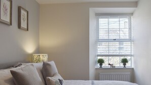 Une chambre à coucher avec un lit, des oreillers, une lampe, une fenêtre avec des stores et deux petites plantes en pot sur le rebord de la fenêtre.