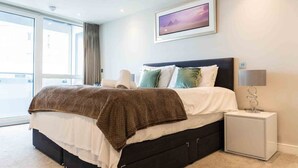 A modern bedroom with a large bed, a bedside table with a lamp, and a view of the outdoors through a large window.
