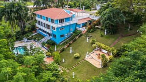 A large, two-story building with a red-tiled roof and a blue exterior, surrounded by lush greenery and a swimming pool. - Your Private Slice of Paradise! (Luquillo)