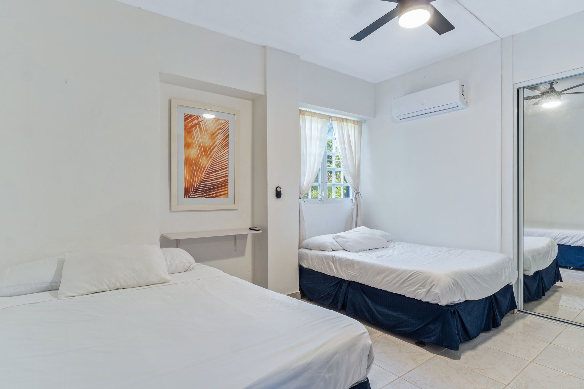 Habitación de hotel con dos camas, ventilador de techo, aire acondicionado, ventana con cortinas e imagen enmarcada en la pared.