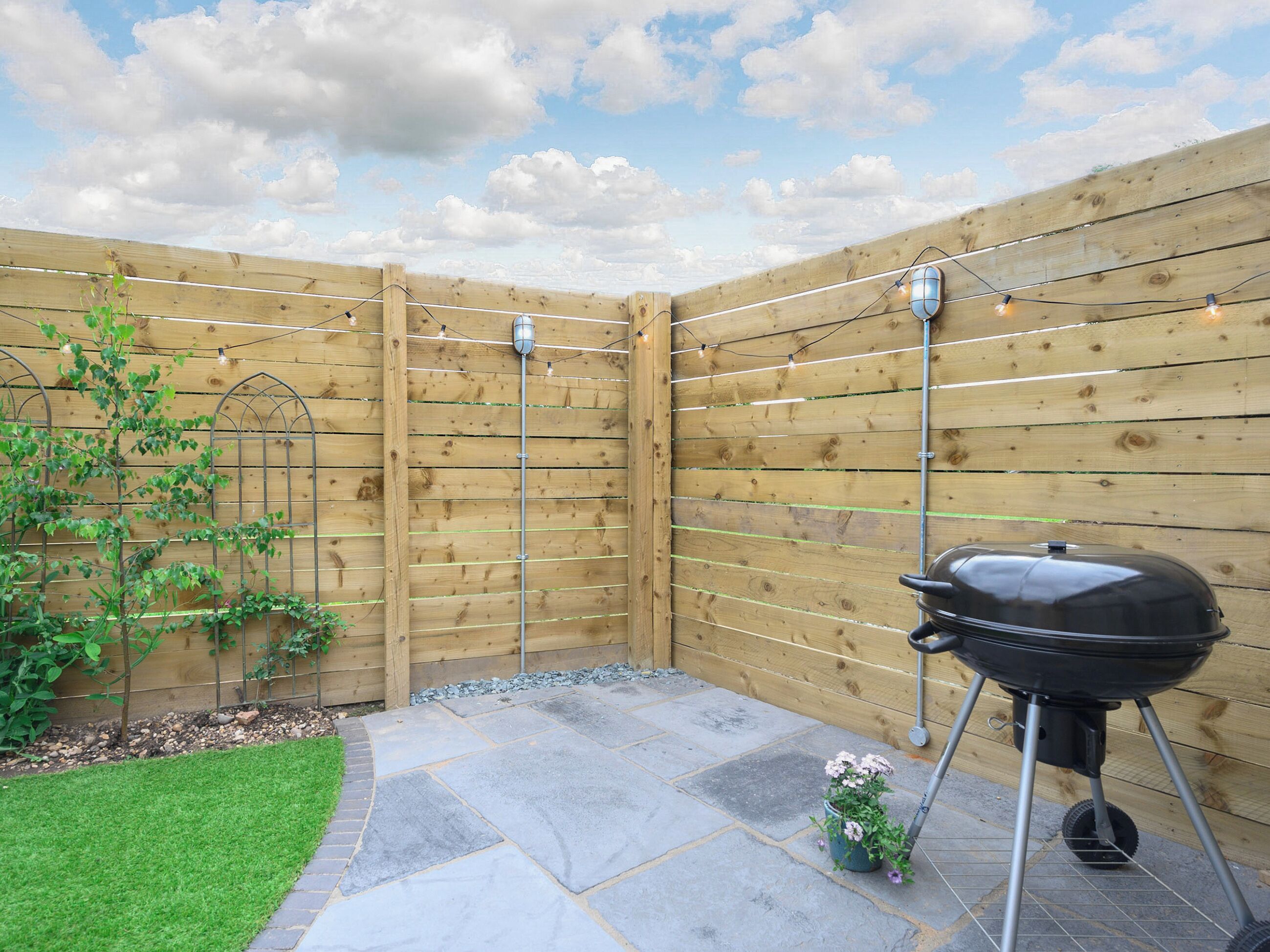 Een achtertuin met houten omheining, een barbecue, sfeerverlichting en een kleine tuin.
