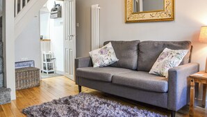 A living room with a grey sofa, floral cushions, a fluffy rug, a wooden side table, and a mirror with an ornate frame. - 1 bedroom accommodation in Keswick (Keswick)