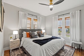 Une chambre moderne avec un grand lit, deux tables de chevet, un ventilateur de plafond et une vue sur l’extérieur.