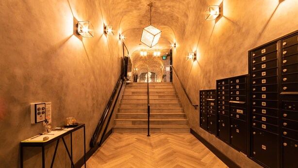 Ein Flur mit Holzdielen im Fischgrätmuster, einer Treppe und einer Reihe schwarzer Spinde rechts.