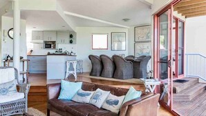 Un amplio salón con un sofá de cuero marrón, un sillón de mimbre blanco y una pequeña mesa blanca. Se ve una cocina con armarios blancos y un microondas al fondo.