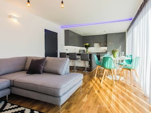 A modern living room with a grey sofa, a dining area with green chairs, and a kitchen in the background.