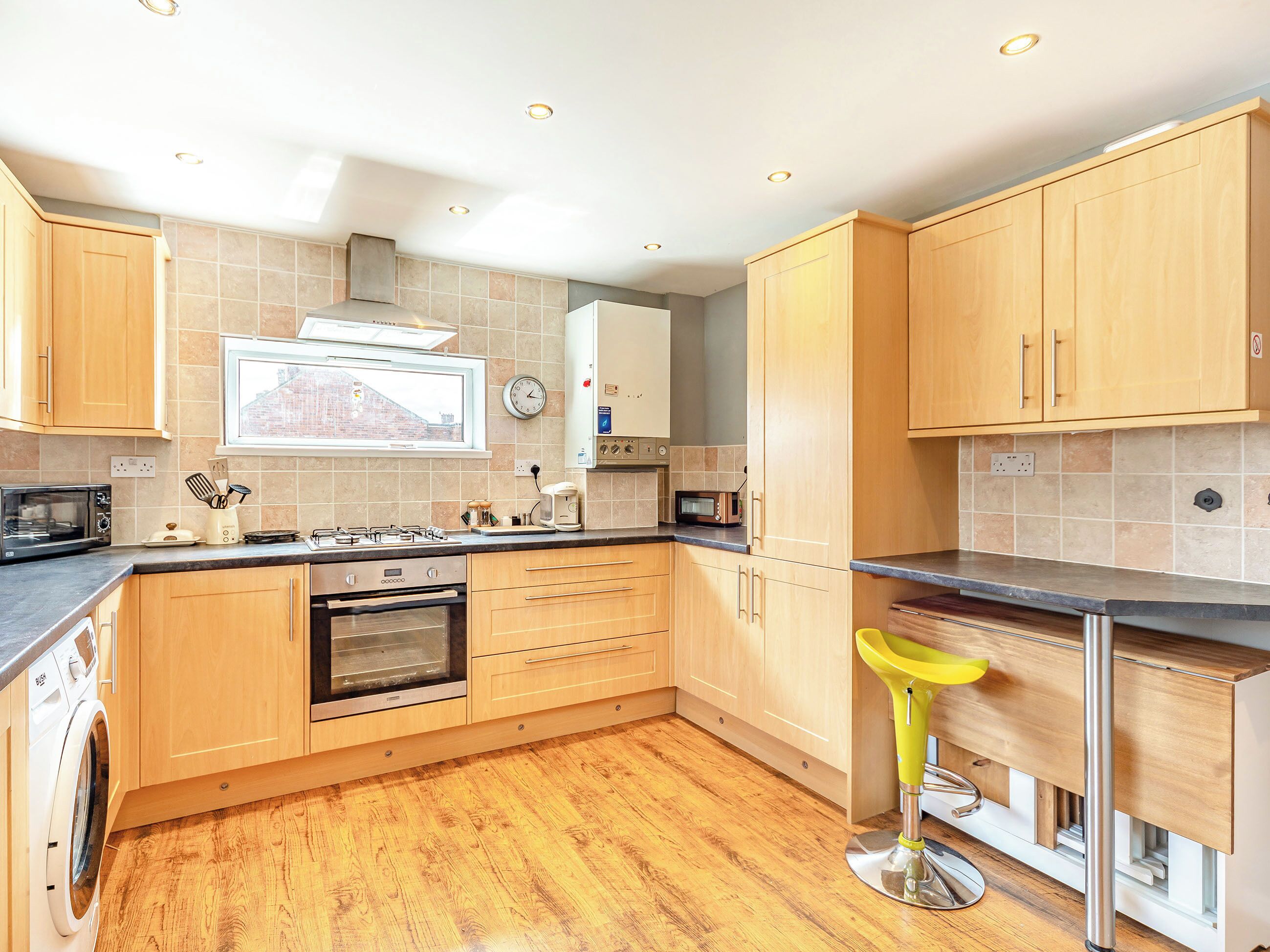 A modern kitchen with wooden cabinets, a built-in oven, a microwave, and a yellow bar stool.