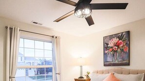 A bedroom with a bed, a ceiling fan, a window with curtains, a nightstand with a lamp, and a painting on the wall. - Colorado Springs Home · Hot Tub · Built for Longer Stays (Colorado Springs)