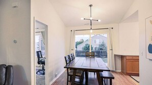 A spacious room with a large wooden dining table, a modern chandelier, and a view of a residential area through large windows. - Colorado Springs Home · Hot Tub · Built for Longer Stays (Colorado Springs)