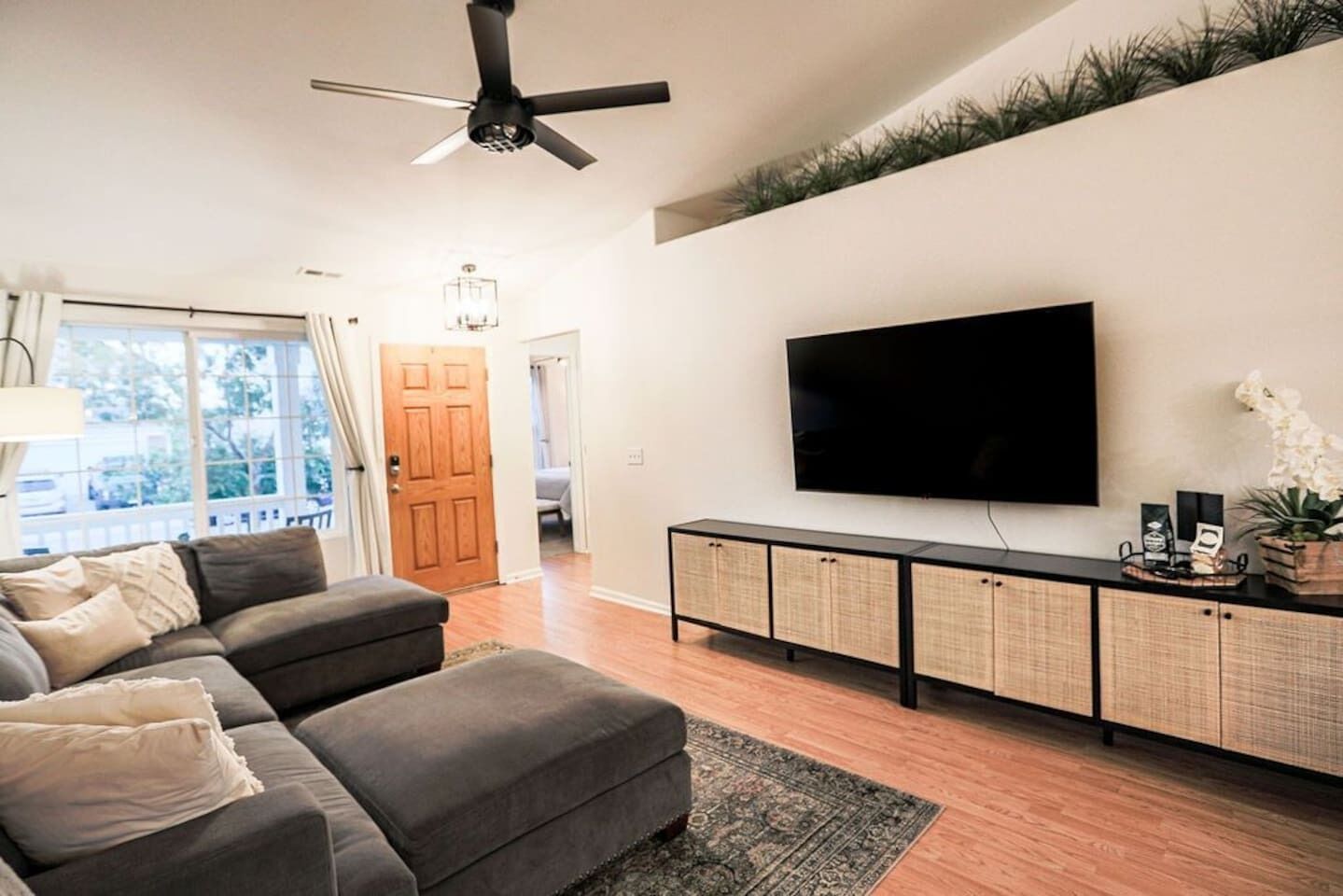 Ein modernes Wohnzimmer mit einem großen Flachbildfernseher, einem dunkelgrauen Ecksofa, einem Holzschrank mit Flechtüren und einem Deckenventilator.