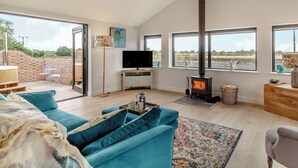 A spacious living room with a large sofa, a television, a fireplace, and a view through large windows.