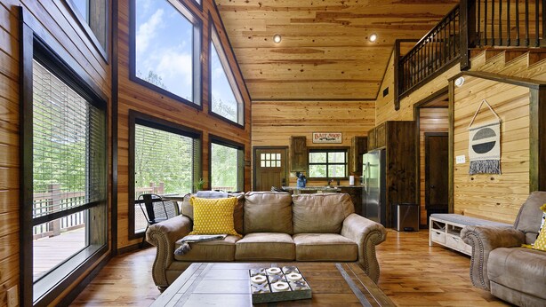 A spacious living room with a wooden ceiling, large windows, a leather sofa, and a coffee table. - Great Views, High Speed Internet, Hot Tub, Fireplace, Balcony, Sauna, Loft! (Broken Bow)