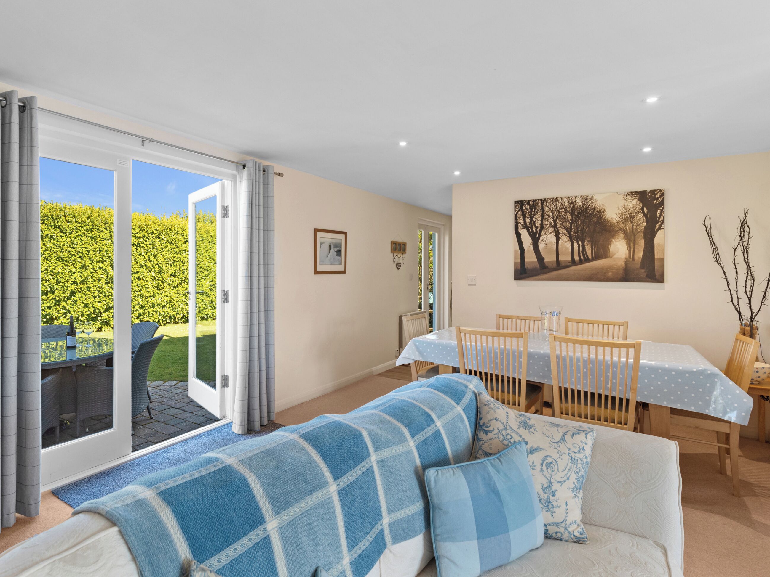 A living room with a sofa, a dining table with chairs, a picture on the wall, and a view of a garden through a sliding door.