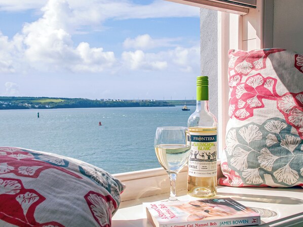 A window view of a body of water with a sailboat, a bottle of wine, a glass, and a book on a windowsill.