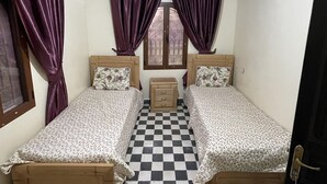 A room with two single beds, a small wooden nightstand, a window with curtains, and a checkered floor. - Dar izniren (Ourika)