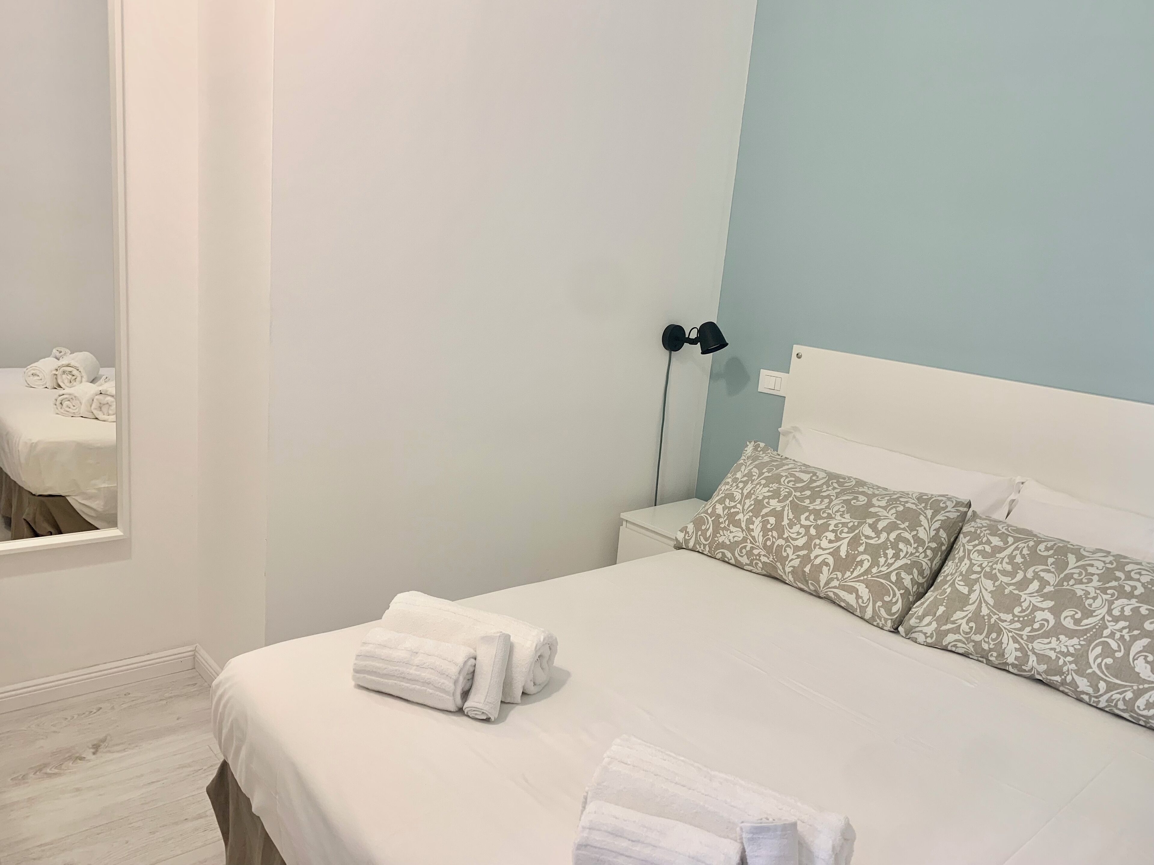 Una cama bien hecha con sábanas blancas y toallas enrolladas, una mesita de noche con lámpara y almohadas estampadas.