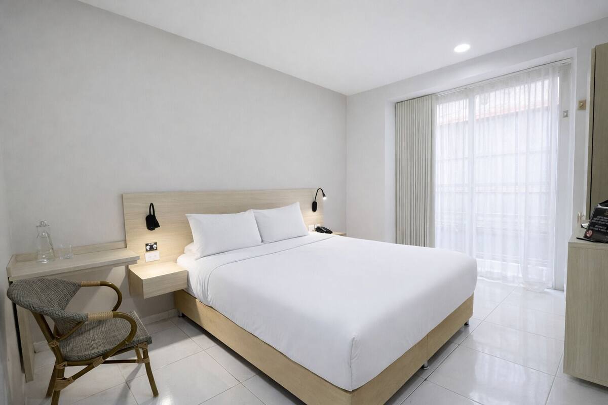 Una habitación de hotel con cama, mesita de noche, lámpara, silla y ventana con cortinas.