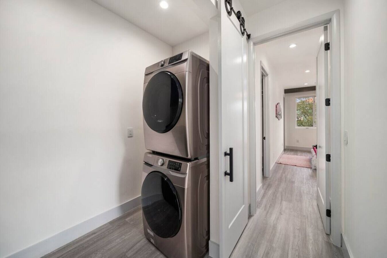 Un lavadero con lavadoras y secadoras de carga frontal apiladas, una puerta blanca y un pasillo que conduce a una habitación con una alfombra rosa.