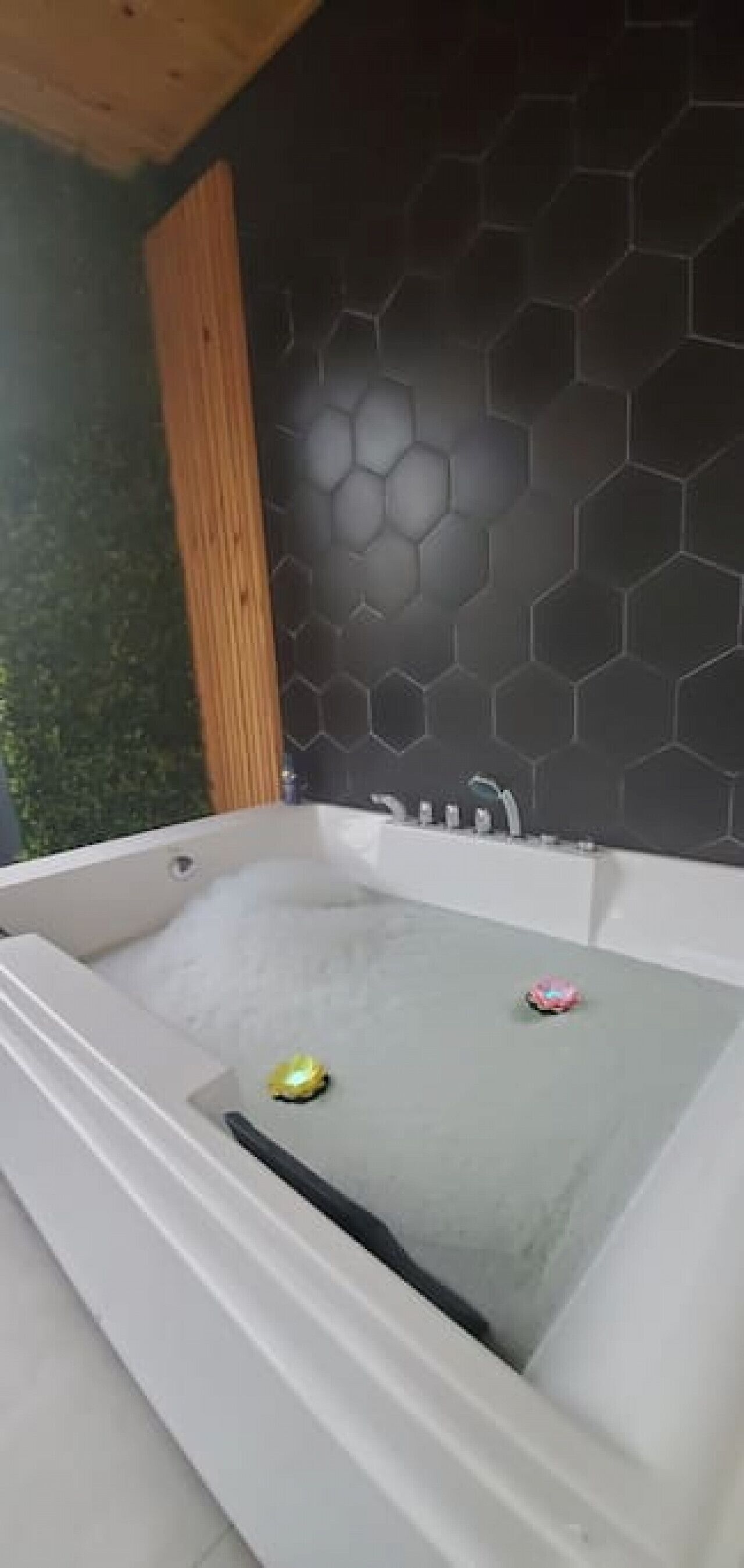A modern bathroom with a large bathtub, a wooden column, and a black hexagonal tile wall.