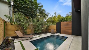 Ein rechteckiger Pool mit einer kleinen Liegefläche, umgeben von einem Holzzaun und üppiger Bepflanzung.