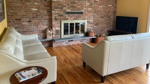 A living room with a brick fireplace, white sofas, a flat-screen TV, and a wooden floor. - Perfect Hamptons Family Fall escape (Hampton Bays)