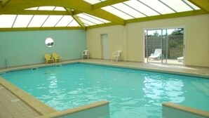 An indoor swimming pool with a tiled floor, yellow ceiling beams, and a view of the outside through a glass door. - Le Cocon des Dunes - Heated swimming pools (Brem-sur-Mer)