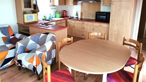 A compact kitchen with wooden cabinets, a microwave, a stove, and a dining area with a table and chairs. - Le Cocon des Dunes - Heated swimming pools (Brem-sur-Mer)
