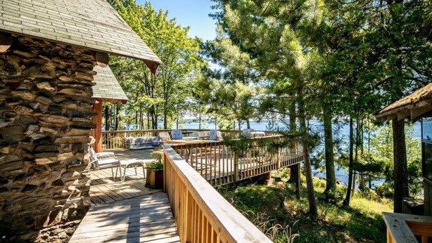 Un bâtiment en pierre doté d’une terrasse en bois qui mène à un quai surplombant l’eau, le tout entouré d’arbres.