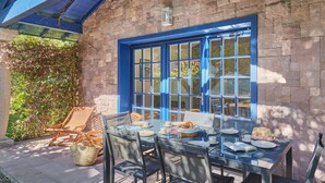 An outdoor dining area with a stone wall, blue-framed windows, and wooden furniture. - Villas Odalys Domaine de Lana (Urrugne)