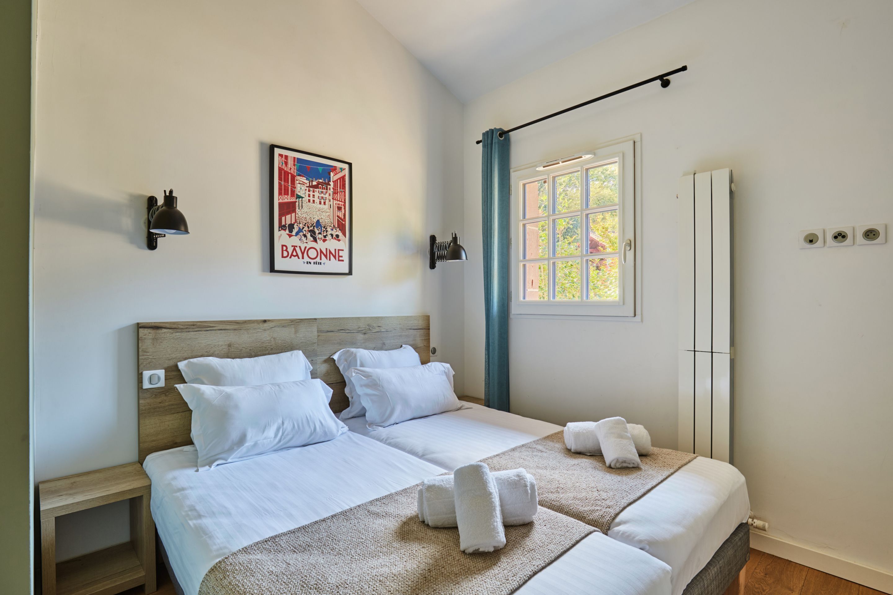 A double bed with white linens and two towels, a wooden headboard, a wall-mounted light, a framed picture, a window with curtains, and a white wardrobe.