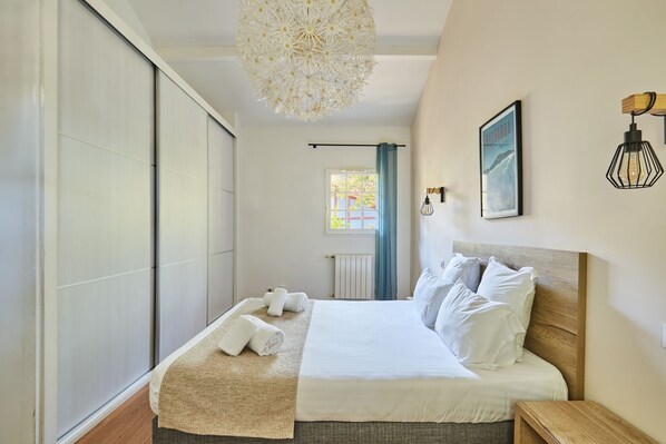 A neatly made bed with white linens and pillows, a woven throw blanket, and a wooden nightstand with a lamp. - Villas Odalys Domaine de Lana (Urrugne)