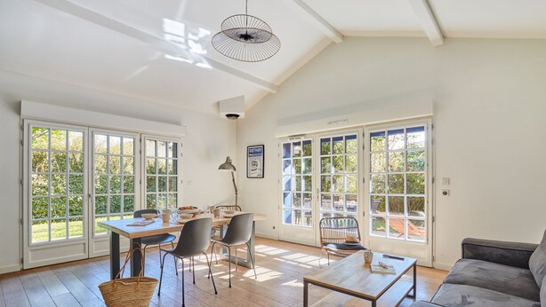 A spacious living room with a wooden floor, a gray sofa, a dining table with chairs, and large windows with a view of greenery. - Villas Odalys Domaine de Lana (Urrugne)
