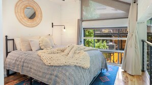 A bedroom with a bed, a ceiling fan, a round mirror, and a window with a view of the outside.