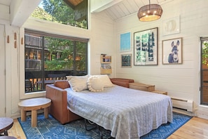 Une chambre confortable avec une grande fenêtre, un lit recouvert d’une literie à motifs, une petite table en bois et un tapis bleu à motifs.