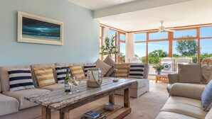 Ein Wohnzimmer mit einer Couch, einem Couchtisch und Blick auf Grün hinter großen Fenstern.