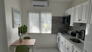 A compact kitchen with white cabinets, a stove, a microwave, and a sink. There's a window with blinds, a green chair, and a plant on the counter. - Sapphire by the Sea Unit 9 - King Pool View Suite King, kitchen, heated pool  (Fort Lauderdale)
