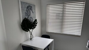 A small dining area with a white table and chairs, a large plant in a vase, a seahorse painting on the wall, and a window with blinds. - Sapphire by the Sea Unit 9 - King Pool View Suite King, kitchen, heated pool  (Fort Lauderdale)