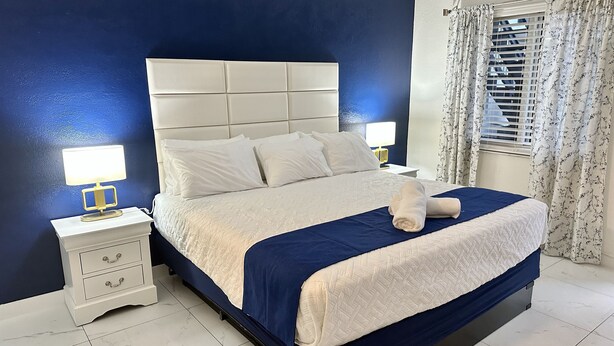 A neatly made bed with a blue bedspread and a white blanket, two bedside tables with lamps, a white nightstand with drawers, and a window with floral curtains. - Sapphire by the Sea Unit 9 - King Pool View Suite King, kitchen, heated pool  (Fort Lauderdale)