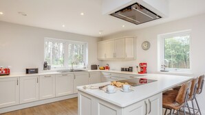 A modern kitchen with white cabinetry, a central island with seating, a built-in oven, and a window with a view of trees. - 4 bedroom accommodation in Craigellachie (Craigellachie)