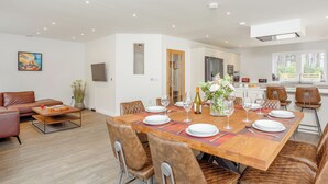 A spacious dining area with a wooden table set for six, surrounded by leather chairs. A kitchen with modern appliances is visible in the background. - 4 bedroom accommodation in Craigellachie (Craigellachie)